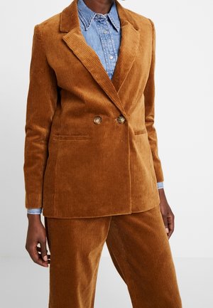 Persoon draagt een bruin corduroy double-breasted blazer en bijpassende broek over een lichtblauw denim overhemd met knopen.