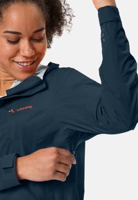 Giacca impermeabile blu navy con tasca con zip, polsini regolabili e fodera interna bianca. Presenta un logo sul petto. Texture liscia.