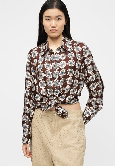 Γυναίκα που φορά καφέ πουκάμισο με κουμπιά και μοτίβο από άσπρες και μπλε μαργαρίτες, δεμένο στη μέση, και μπεζ παντελόνι με μικρές τρουκς.