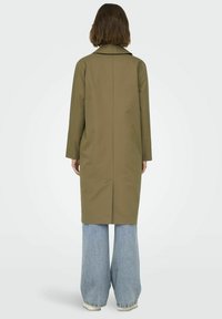 Cappotto verde oliva di media lunghezza con colletto e maniche lunghe; linea dritta con cucitura sul retro; indossato sopra jeans in denim blu a gamba larga.