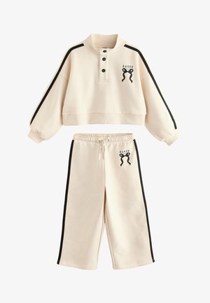 Beige cropped sweatshirt med lange ærmer og bukser med vide ben, med sorte sidelister og sløjfe-logo detalje på brystet og låret.