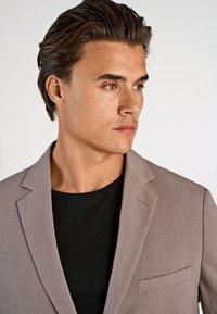 Jasnoszara marynarka z notched klapami, pojedynczą kieszenią na piersi i dopasowanym krojem, noszona na czarnej koszulce, o gładkiej fakturze.