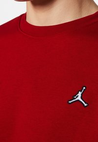 T-shirt rouge à col rond porté par une personne, avec un petit logo Jumpman blanc brodé sur le côté gauche de la poitrine.