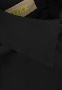 Collare di camicia nera con una texture liscia e opaca. L'etichetta interna presenta 'IJXX' in giallo brillante su uno sfondo beige.