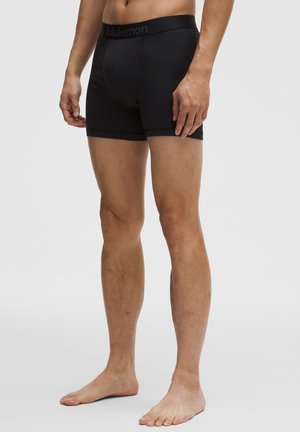 Hombre con bóxers negros de Lululemon de pie descalzo contra un fondo liso.