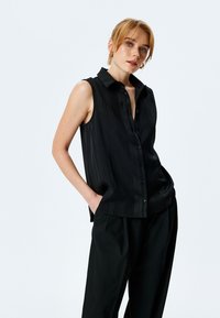 Maglietta nera senza maniche con righe verticali, design con colletto, chiusura con bottoni frontale e vestibilità comoda, abbinata a pantaloni neri larghi.