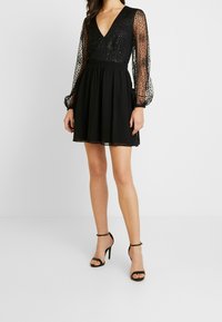 Zwarte jurk met een diepe V-hals en doorzichtige lange mouwen met stippen, met een getailleerde bodice en een uitlopende rok. G gedragen met zwarte sandalen met banden.