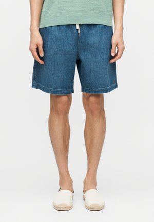 Mann trägt blau-gefärbte Jeans-Shorts mittlerer Länge, hellgrünes T-Shirt und weiße Espadrilles zum Hineinschlüpfen, steht vor einem einfarbigen weißen Hintergrund.