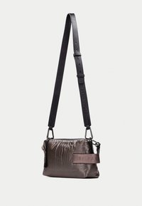 Sac en nylon rembourré en gris foncé avec une sangle texturée noire, comportant des accents métalliques et un patch logo décoratif.