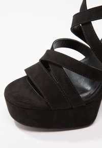 Sandale plateforme en suède noir avec plusieurs larges brides croisant le pied et une semelle épaisse, inclinée pour montrer les détails de face et de côté.