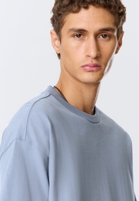 Světle modrá mikina s kulatým výstřihem, volným střihem, texturou látky a mírně spadlými ramenními švy. Bez výrazných vzorů nebo akcentů.