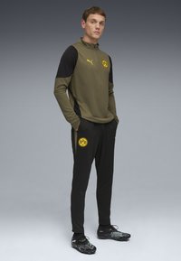 Olivengrøn atletisk top med sorte ærmer, der har et logo. Sorte bukser med logo, parret med sorte og grønne støvler. Glat tekstur.