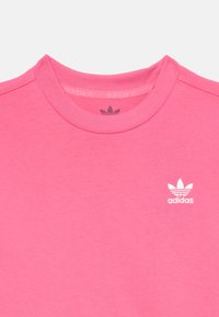Roza bombažna majica s krožnim izrezom in belim logotipom Adidas na prsih. Gladka tekstura in preprost dizajn, primerna za vsakodnevno nošenje.