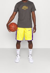 Persona vestida con una camiseta gris de los Lakers, pantalones cortos de baloncesto amarillos y morados, calcetines y zapatillas Nike blancas, sosteniendo un balón de baloncesto naranja a su lado.