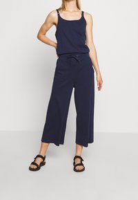 Vrouw in marineblauw mouwloos topje en wijde cropped broek met trekkoord in de taille, gecombineerd met zwarte sandalen, staand tegen een effen achtergrond.