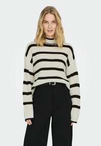 Gestreifter Strickpullover in Creme und Schwarz, mit hohem Kragen und langen Ärmeln. Über einer schwarzen, hoch taillierten Hose getragen.