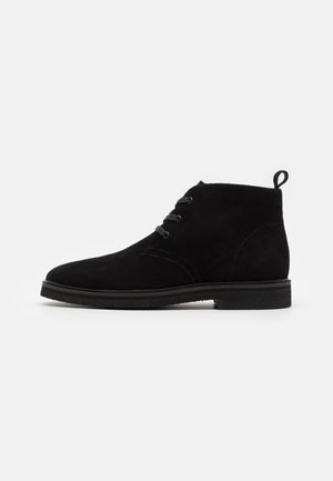 Stivaletti in suede nero con punta arrotondata, due occhielli per i lacci e una piccola linguetta sul retro. Suola in gomma piatta.