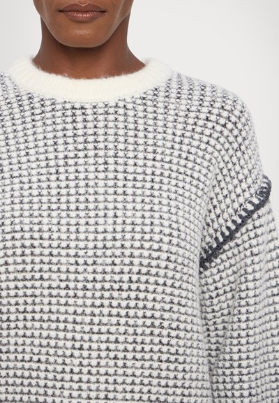 Knitted sweater with a white and navy grid pattern, featuring a ribbed crew neck and textured fabric with distinct stitching details.
