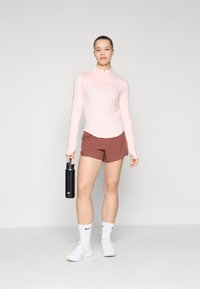 Lys rosa langermet treningstopp med glidelås og brune shorts, hvite sokker og hvite sko. Holder en svart vannflaske.