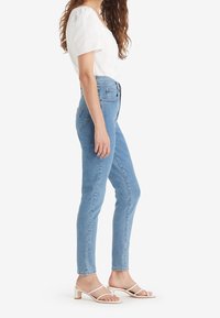 Mujer de pie de lado con una blusa blanca de manga corta, unos jeans ajustados de talle alto color azul claro y sandalias de tacón blancas.