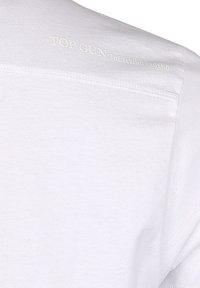 Vit bomull t-shirt med rund hals och korta ärmar, med präglad text "TOP GUN THE FLYING LEGEND" på baksidan nära kragen.