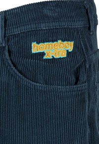 Tumši zila korduroja auduma virsma ar vertikālām svītrām, ar izšūtu "homeboy x-tra" logo spilgti oranžā un zilā krāsā uz kabatas zonas.
