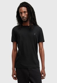 Uomo con lunghi dreadlocks che indossa una maglietta nera a maniche corte con un piccolo logo di un animale cornuto bianco sul petto, in piedi su uno sfondo semplice.