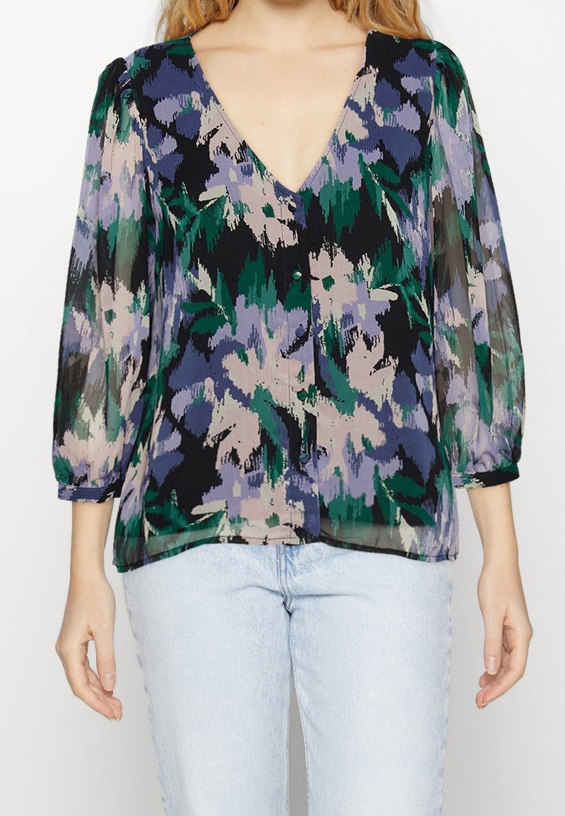 Femme portant un chemisier floral semi-transparent aux teintes bleues, vertes et violettes avec des manches trois-quarts bouffantes, associé à un jean bleu clair.
