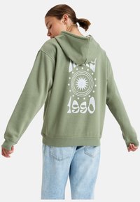 Sudadera verde oliva con un diseño gráfico blanco en la parte posterior, que presenta un motivo de sol y el año "1990". Tejido suave y texturizado con un corte relajado.