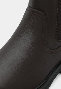 Brown leather ankle boot featuring a textured finish, contrasting stitching, and elastic side panels for ease of wear. Black rubber sole.