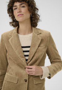 Blazer en velours côtelé couleur beige clair avec revers cranté, fermeture à bouton unique et poches avant, superposé sur un pull rayé beige et noir.