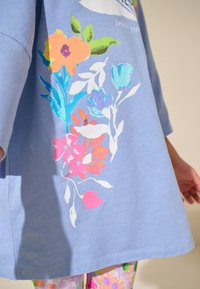 Világoskék rövid ujjú felső, amely élénk rózsaszín, narancssárga és kék virágmintákkal díszített az elején, laza szabású és sima tapintású.