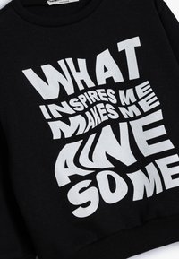 Zwarte sweatshirt met een gedurfde, vervormde witte tekst: "WAT MIJ INSPIREREND IS MAAKT ME LEVEND SOME." Zachte stof met geribbelde manchetten.