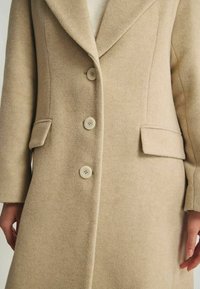 Manteau en mélange de laine beige avec un col cranté, fermeture à trois boutons, deux poches latérales et une texture lisse.