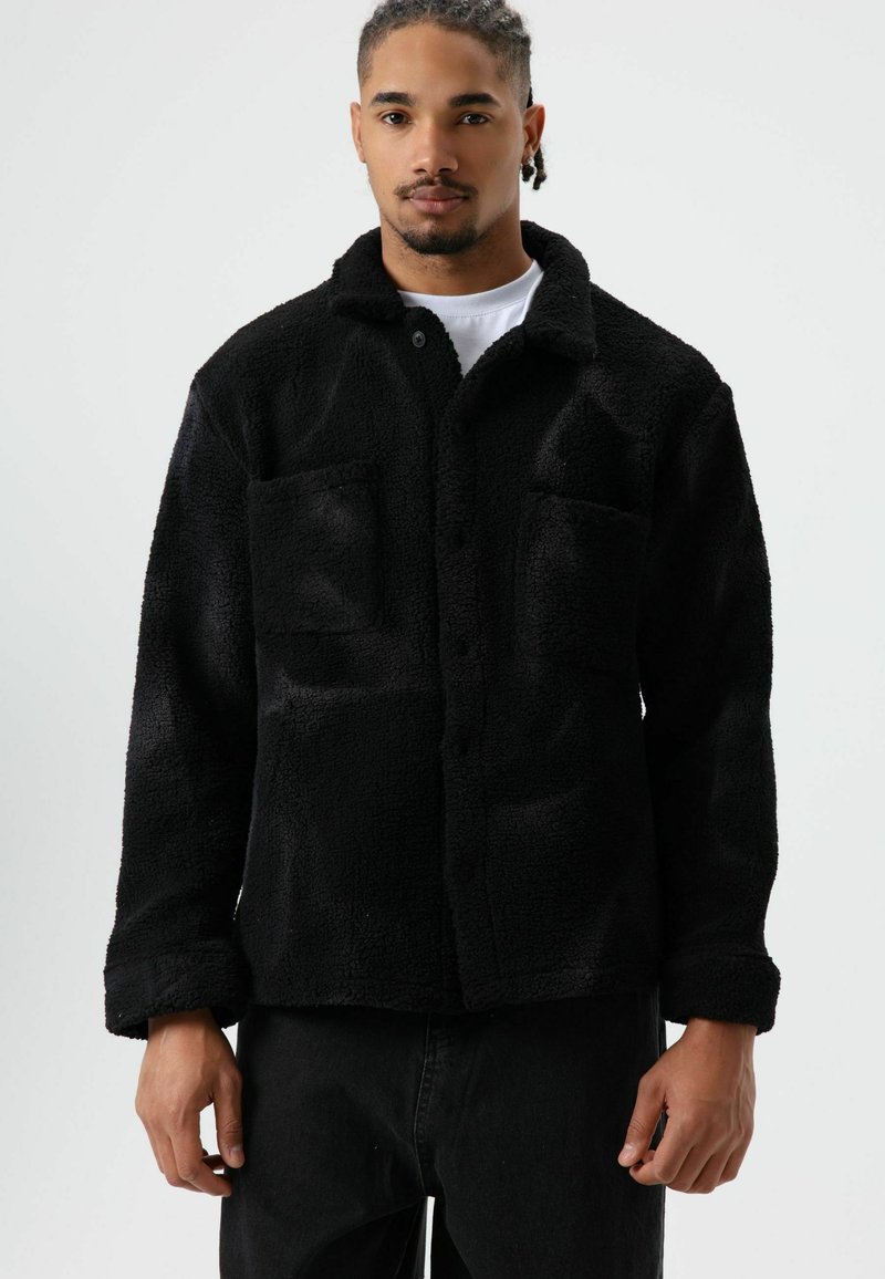 Zwarte fleecejas met een getextureerd oppervlak, voorzien van een kraag, knoopsluiting aan de voorkant en twee borstzakken. Eenvoudig, casual ontwerp.