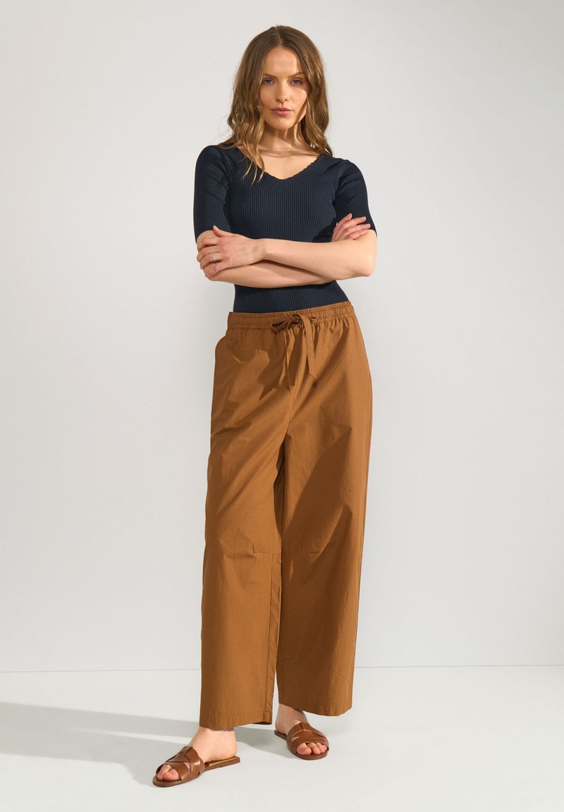 Femme debout, bras croisés, vêtue d'un top côtelé noir, d'un pantalon marron ample avec cordon et de sandales marron à enfiler, sur un fond uni.