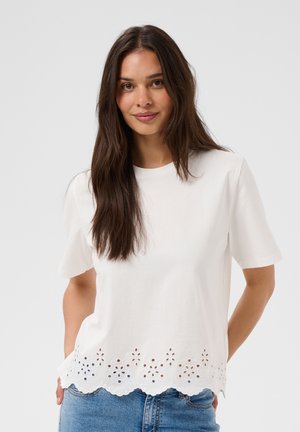 Mujer joven con el pelo largo y castaño, que lleva una camiseta blanca de manga corta con dobladillo recortado con diseño floral y vaqueros azules, de pie contra un fondo liso.