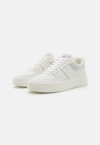 White sneakers with a textured leather upper, perforated detailing, a padded tongue, and a thick, flat rubber sole. Accents in gold.