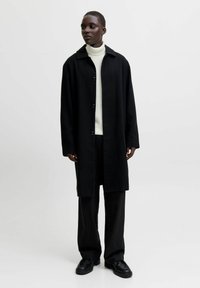 Abrigo de lana negro con cuello solapa, cierre de botones y mangas largas, llevado sobre un suéter de cuello alto blanco y pantalones negros a rayas.