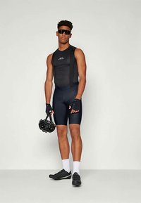 Vyras, vilkintis juodus dviratininko šortus, be rankovių džersį, pirštines, baltas kojines, juodus batus ir akinius nuo saulės, laiko juodą dviratininko šalmą.