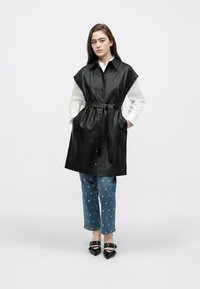 Black leather sleeveless coat with a belted waist, paired with blue denim jeans featuring white circular patterns. Flat black shoes.