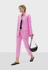 Pink blazer and matching trousers made of smooth fabric; paired with a black top and small black handbag. Footwear features pointed toes.
