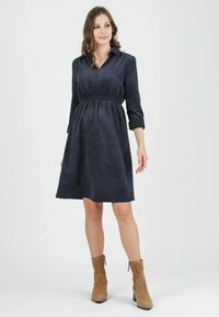 Marineblaues Cordkleid mit kragenförmigem Design, tailliertem Schnitt und Dreiviertelärmeln. Kombiniert mit beigefarbenen Stiefeletten. Geschmeidige Textur.