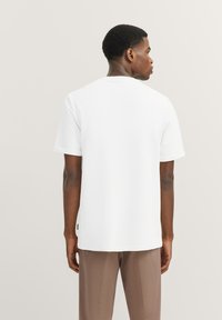 Weißes T-Shirt mit kurzen Ärmeln, lockerer Passform, glatter Textur und hochgekrempelten Ärmeln, kombiniert mit braunen Hosen. Kleines Markenetikett sichtbar.
