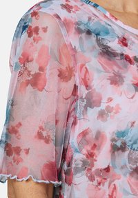Transparente Bluse mit floralem Muster, aus pinkem Stoff mit blauen und roten Blumenmustern, kurzen Ärmeln und einem gewellten Saum.
