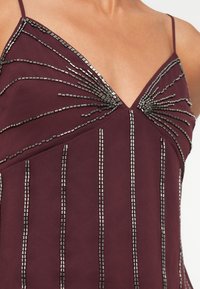 Bordo obleka z nastavljivimi trakovi, ki vsebuje sijoče, linearne okraske iz perl in V-izrez za prilegajoč silhueto.