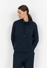 Marineblaues Sweatshirt mit Trichterkragen und Kordelzug, versehen mit dem bedruckten Text "ADVENTURES" auf der Vorderseite, kombiniert mit passenden Hosen.