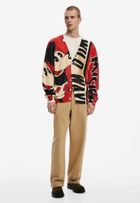 Knitted cardigan in red, cream, and black featuring a cartoon graphic and text. Buttons down the front, relaxed fit, paired with beige pants.
