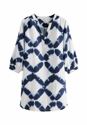 Femme portant une robe ample à manches longues avec des motifs tie-dye noirs et blancs en forme de losanges et un décolleté en V.