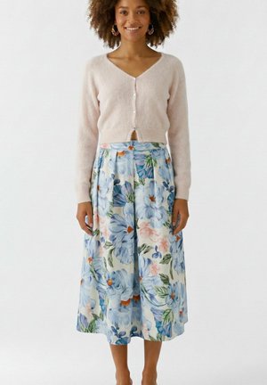 Frau trägt eine hellrosa geknöpfte Strickjacke und einen hoch taillierten Midirock mit blauem Blumenmuster auf weißem Hintergrund.